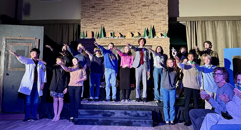 A group of high school aged kids raising their hands on a stage. They appear to be actors in a play.