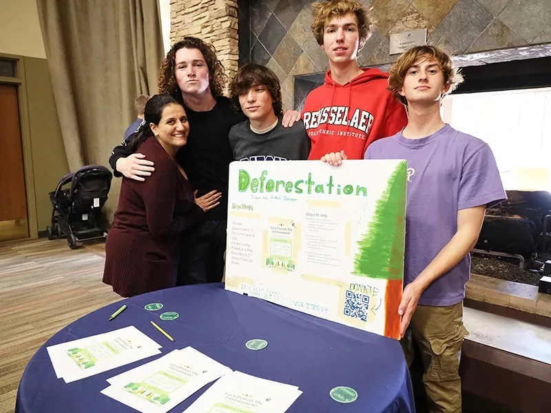 Image showing a group of four male high school students with a female teacher at a table. The students are holding a poster that says "Deforestation" with information on it.