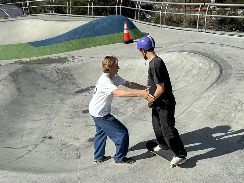A high school boy holding hands with another boy on a skateboard in a skate park to help him balance