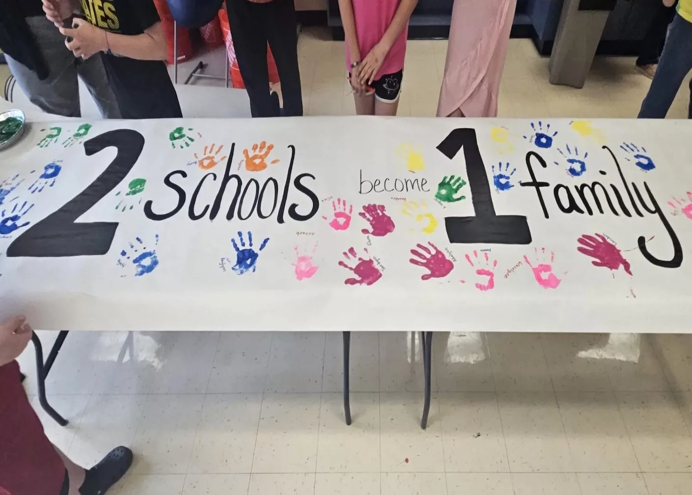 A paper sign with painted handprints with text that reads "2 schools 1 family"