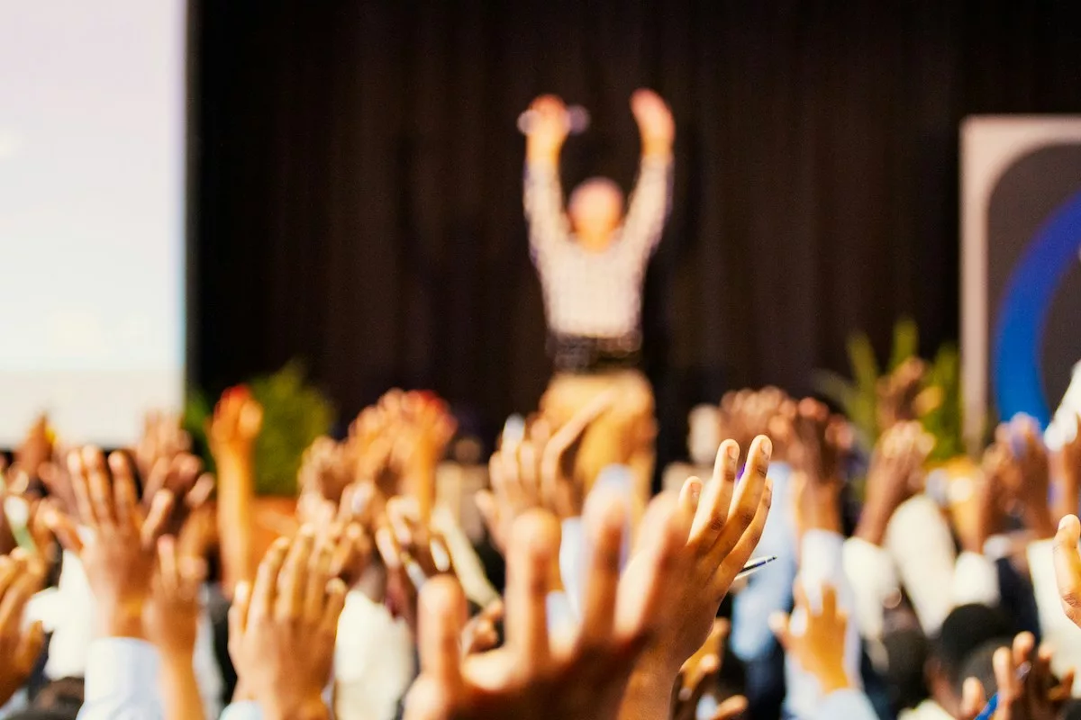 A speaker and audience members raising both their hands at a conference