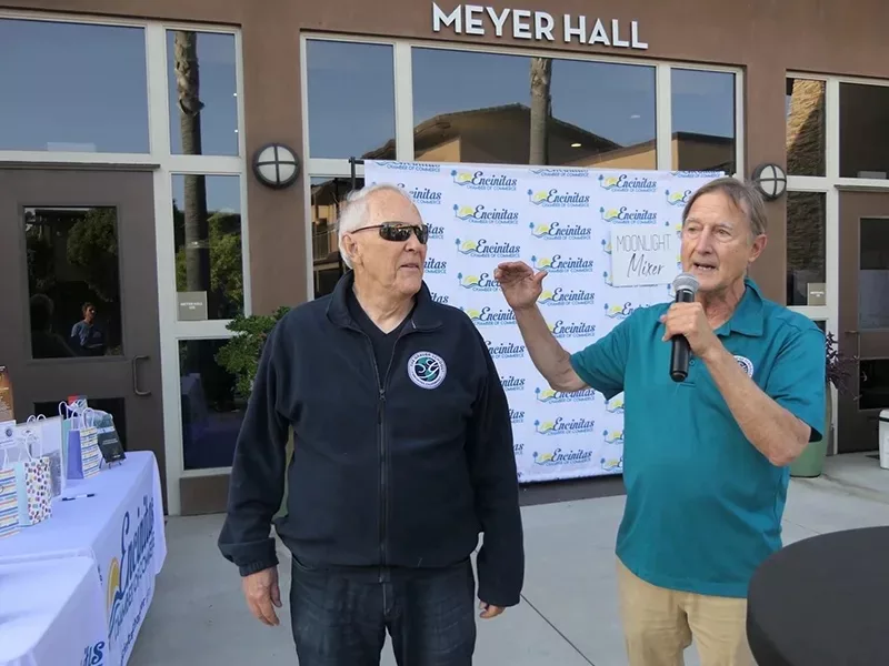 Stuart Grauer speaking in a microphone with an older man next to him in front of a brick building with windows