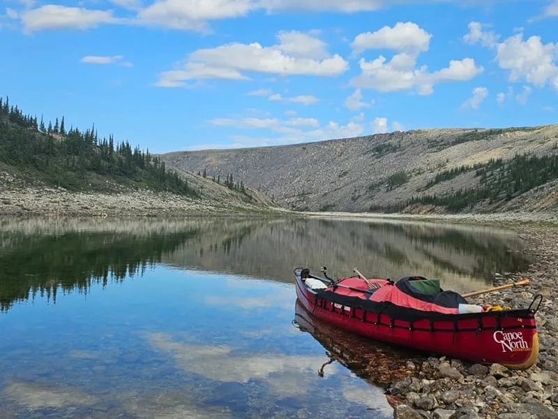 A beautiful view of the Arctic with a red canoe resting on shore and mountains with water and clouds behind it