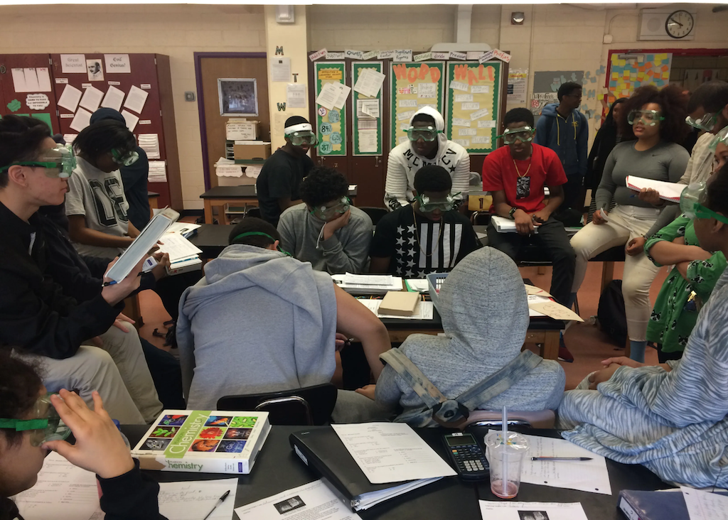 Eleventh-grade students at Urban Assembly Gateway for Technology gather around a lab table, preparing beakers and wearing safety goggles before a chemistry experiment.